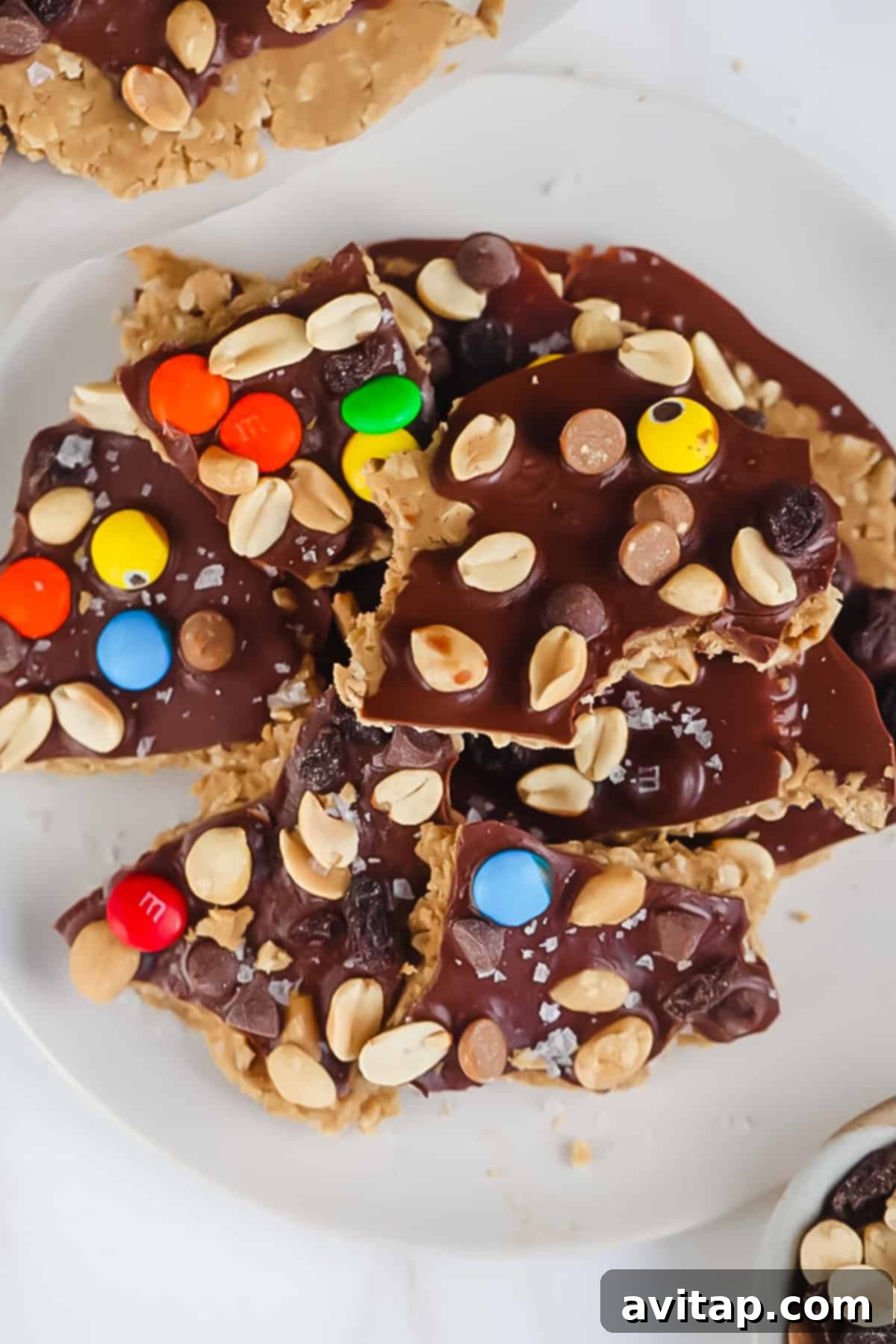 A plate of peanut butter and chocolate bark topped with colorful candy-coated chocolates, peanuts, and sea salt flakes, on a white surface.