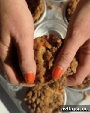 Hands delicately splitting a freshly baked cottage cheese carrot cake muffin, revealing its soft, fluffy interior with visible specks of carrot. A tray of muffins rests in the background.
