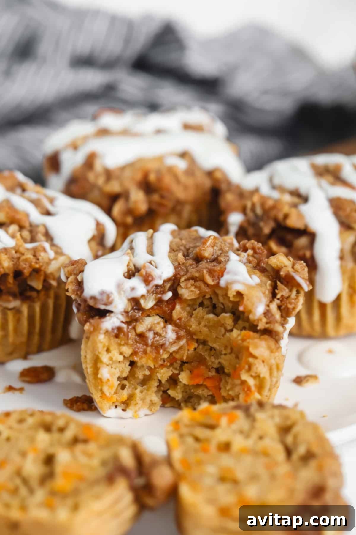 A close-up shot of a plate of Cottage Cheese Carrot Cake Muffins, featuring a crumbly streusel topping and a generous drizzle of white icing. One muffin is partially eaten, showing its moist, tender interior.