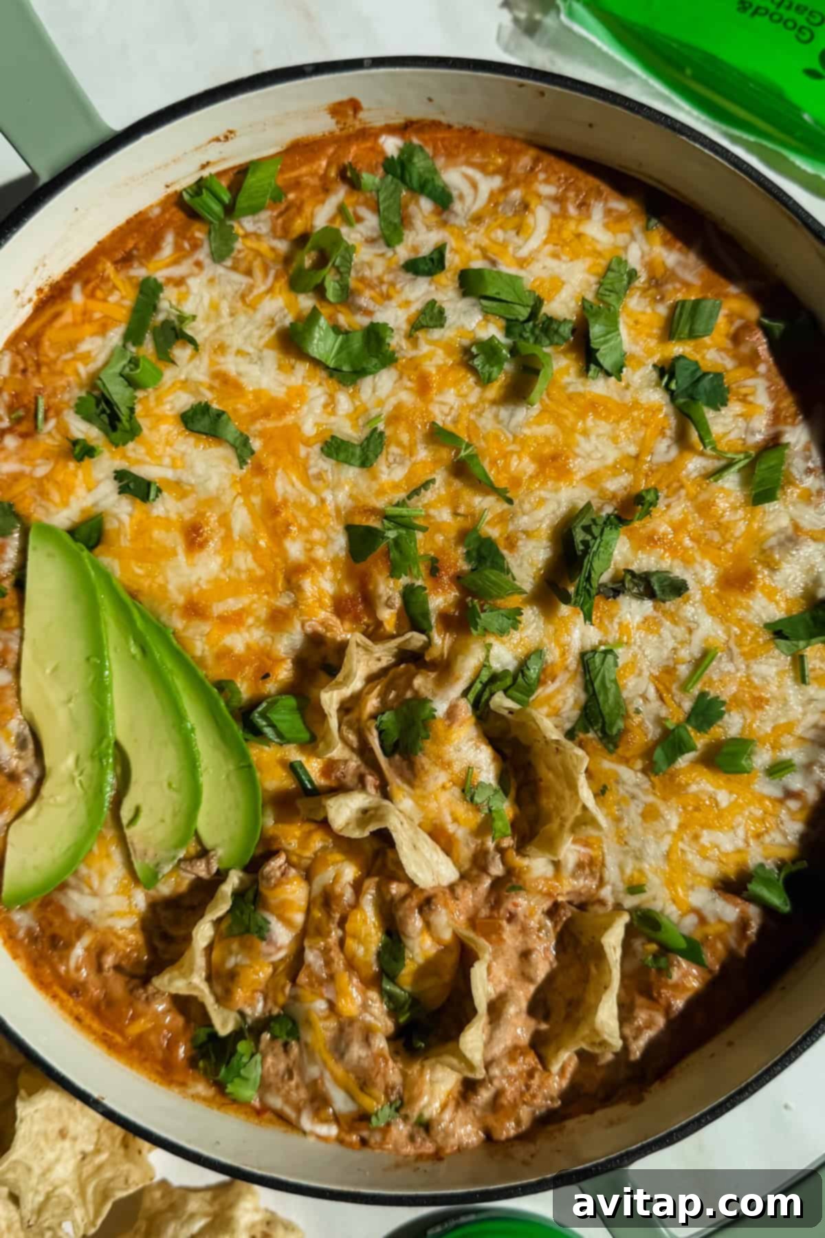 A vibrant dish of cheesy ground beef cottage cheese queso dip, generously garnished with fresh cilantro and avocado slices, surrounded by crispy tortilla chips in a white casserole dish.