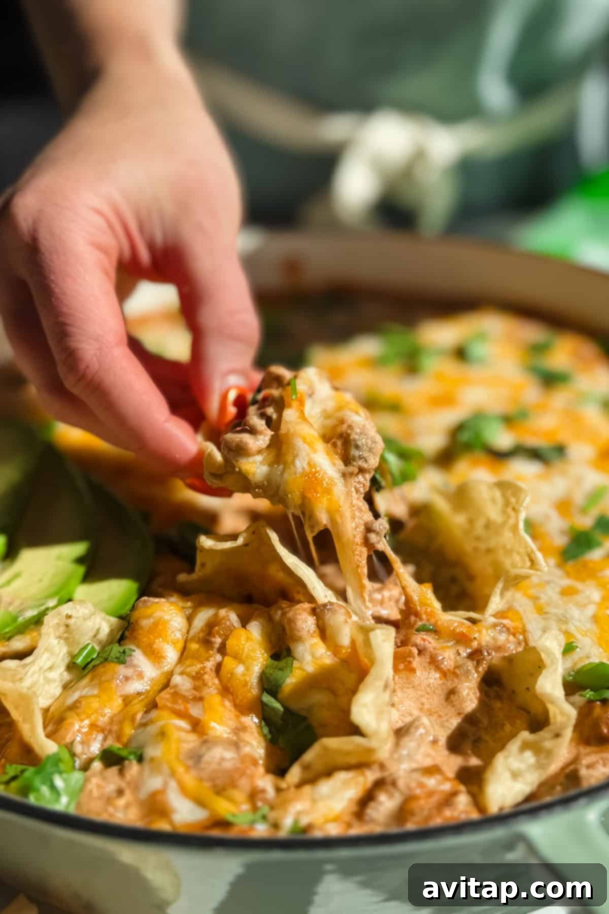 A hand reaching for a serving of hot, cheesy ground beef cottage cheese queso dip, garnished with fresh cilantro and sliced avocado, with a side of crunchy tortilla chips.