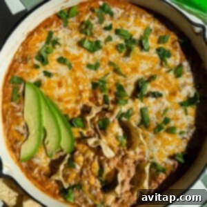 A baked dish of ground beef cottage cheese queso, topped with melted cheese, garnished with sliced avocado and fresh herbs, next to tortilla chips.