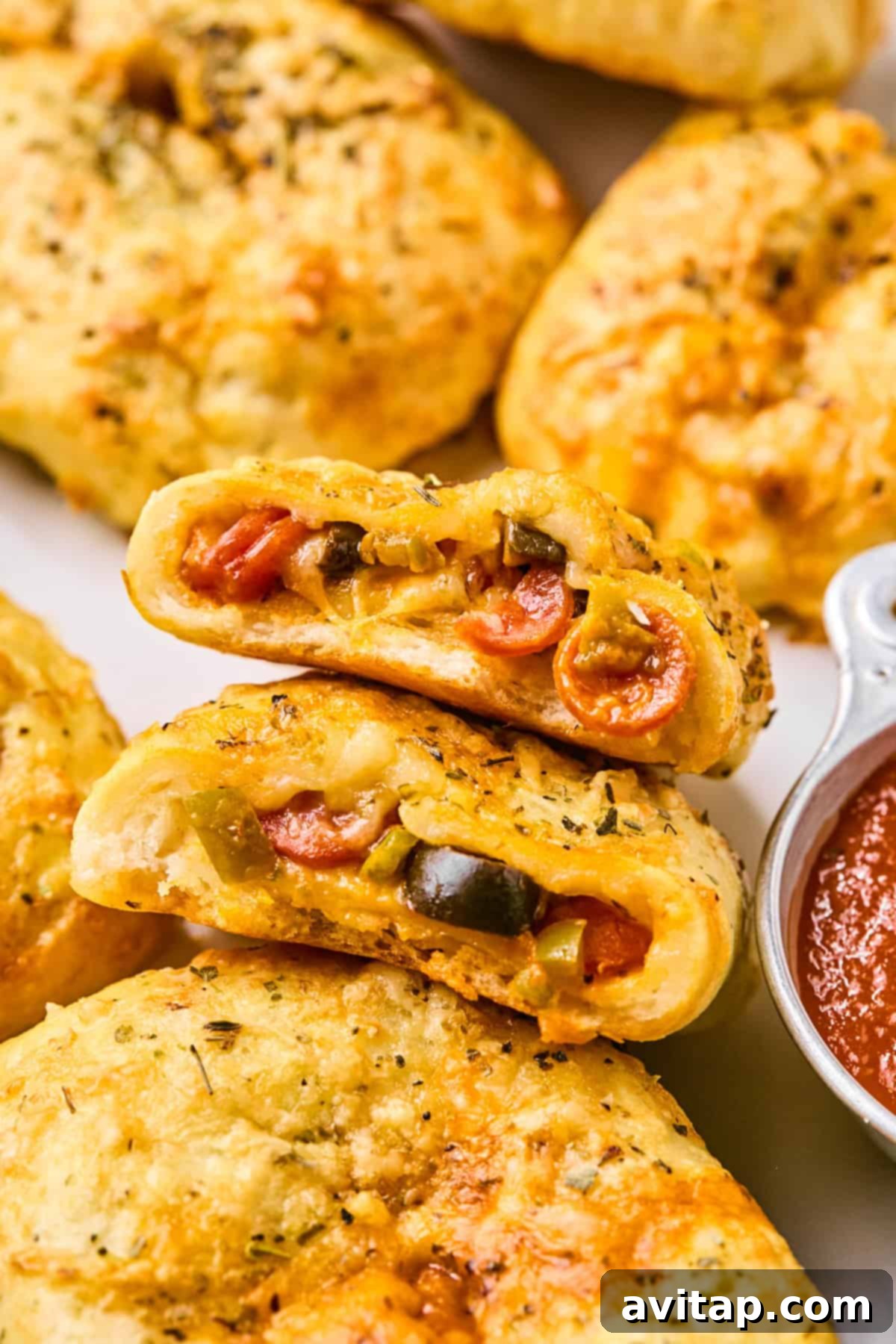Close-up of baked calzones, reminiscent of pizza hot pockets, filled with cheese, pepperoni, and vegetables, accompanied by a small dish of marinara sauce.