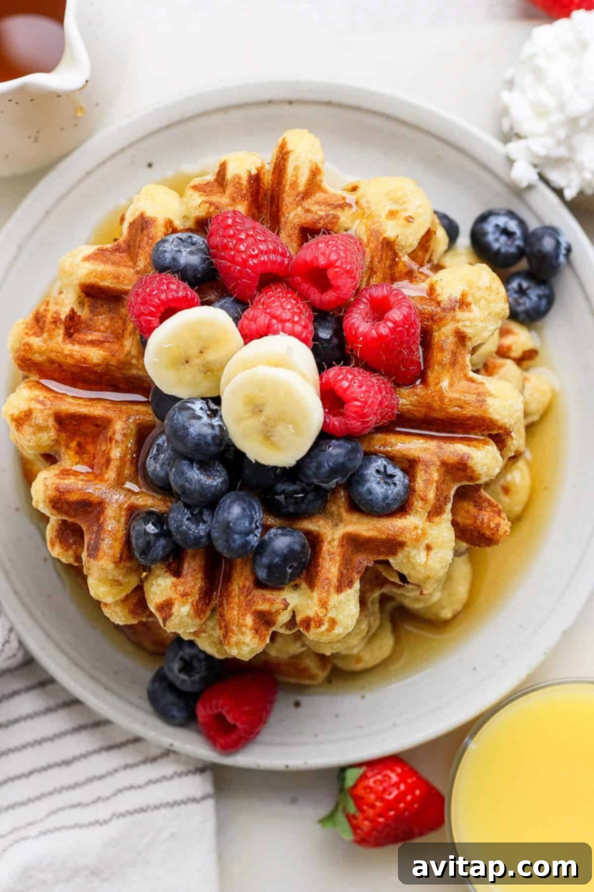 A stack of golden-brown cottage cheese waffles, generously topped with fresh blueberries, raspberries, and slices of banana on a pristine white plate. A glass of refreshing orange juice and a striped napkin sit elegantly nearby, completing the perfect breakfast scene.