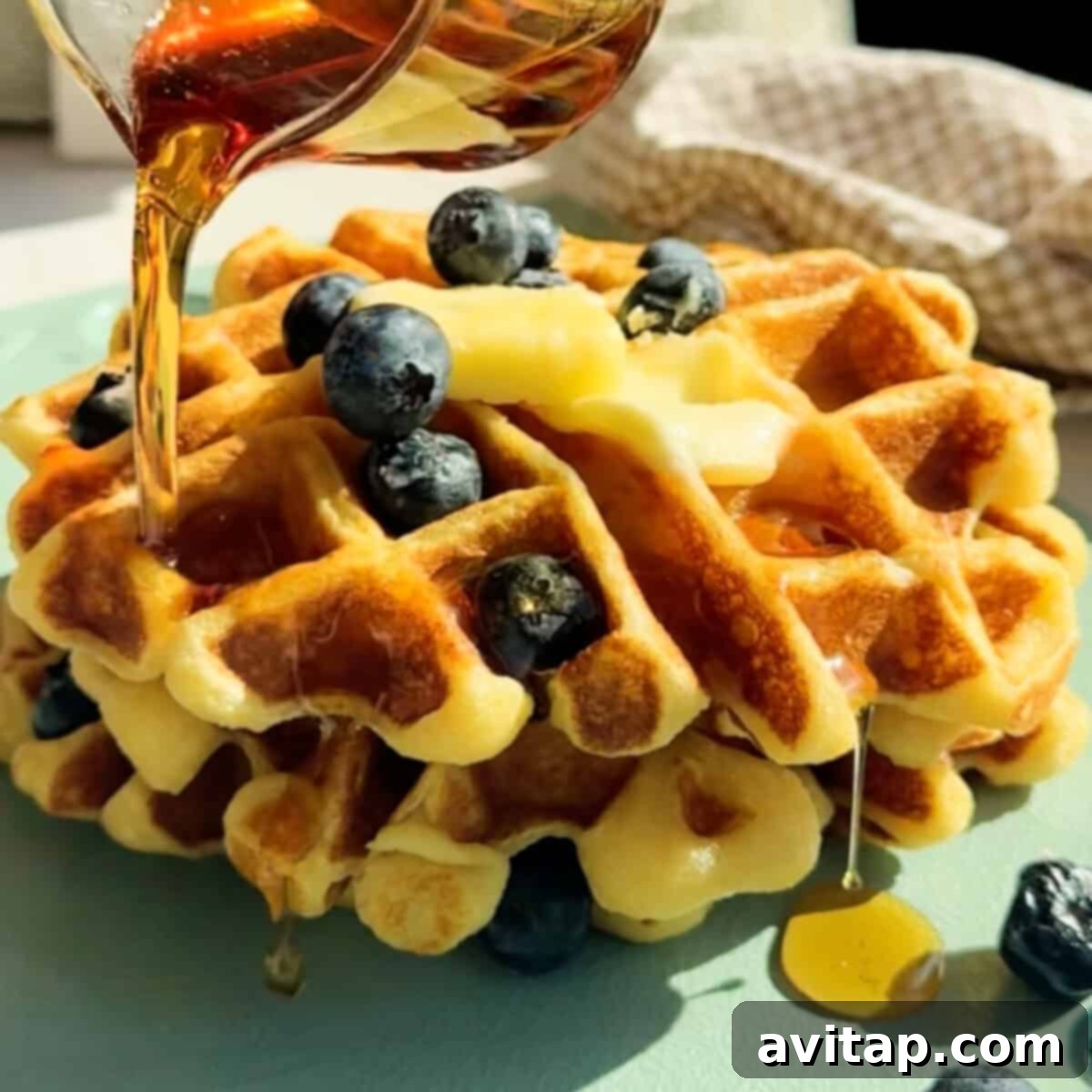High-Protein Cottage Cheese Waffles 3 A close-up shot of golden cottage cheese waffles on a light green plate, generously adorned with fresh blueberries and melting butter, with a stream of golden syrup being drizzled over them.
