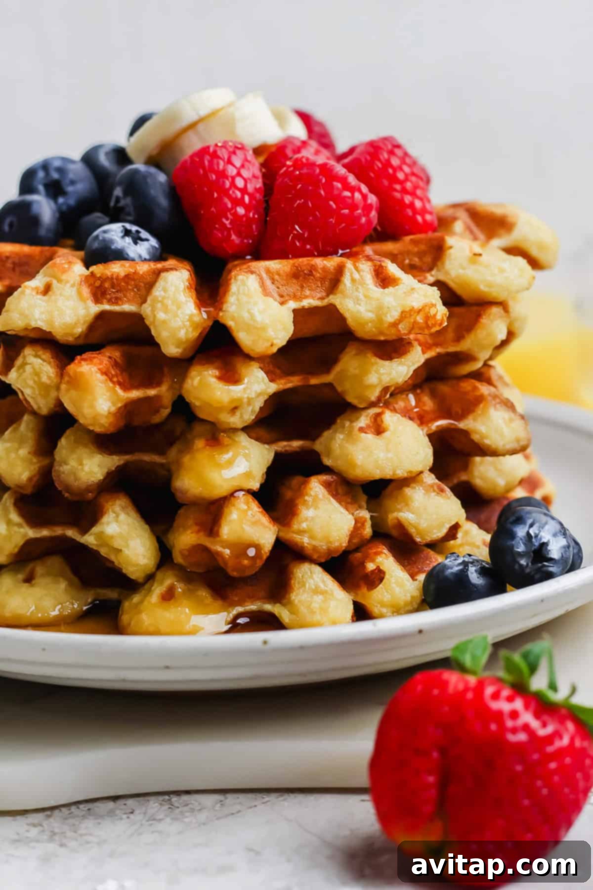 High-Protein Cottage Cheese Waffles 5 A beautifully arranged stack of golden cottage cheese waffles, generously adorned with fresh banana slices, bright red raspberries, and plump blueberries, all artfully drizzled with sweet maple syrup. A single, ripe strawberry rests on the edge of the plate.