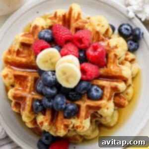 A stack of golden brown cottage cheese waffles, beautifully topped with fresh blueberries, raspberries, and banana slices, all drizzled with a generous amount of syrup on a round white plate. A strawberry adds a touch of color on the side.