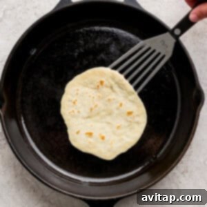 A hand uses a spatula to lift a perfectly cooked, bubbly protein tortilla from a hot black cast iron skillet on a light-colored surface.