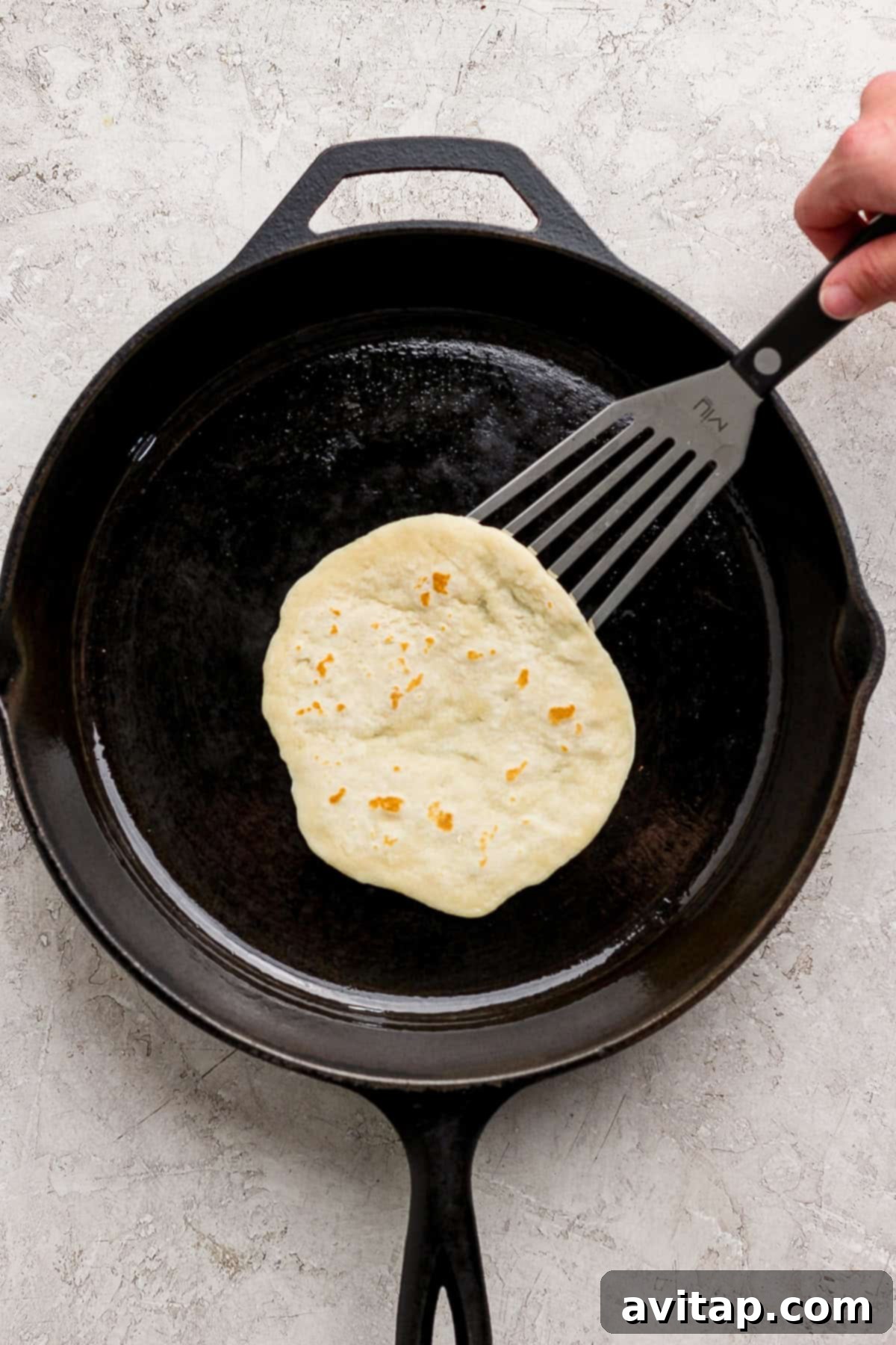 A hand expertly uses a spatula to lift a homemade protein tortilla cooking and bubbling in a hot black cast iron skillet on a clean light-colored countertop.