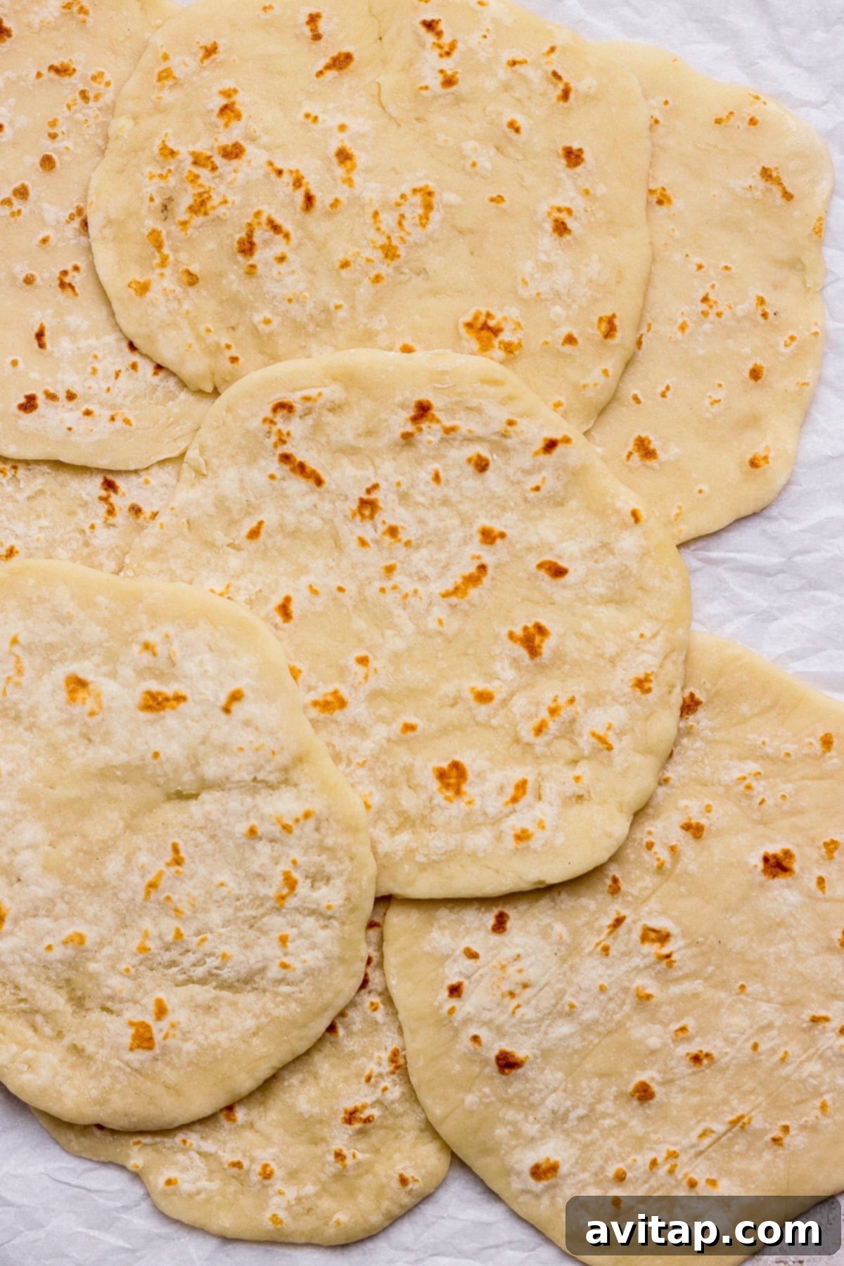 Several soft, slightly browned homemade protein flatbreads, artfully overlapping, presented on a crumpled white textured surface, showcasing their delicious texture.