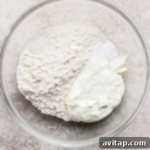 A glass bowl containing flour, salt, and a dollop of plain yogurt, ready to be mixed, on a light textured surface.