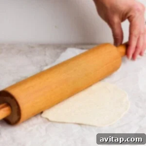 A hand holds a wooden rolling pin, flattening a piece of dough on parchment paper on a light-colored kitchen surface, creating a tortilla.