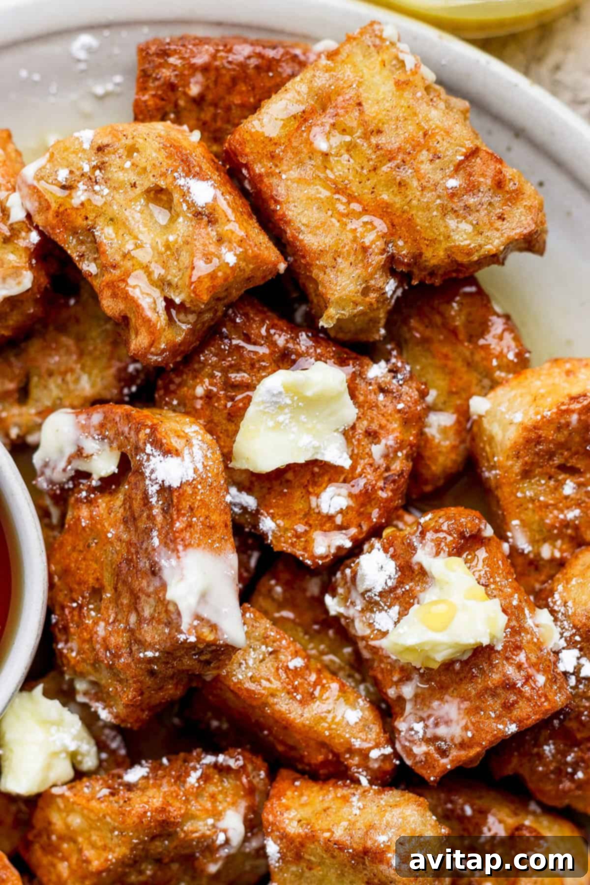 Close-up of golden French toast bites topped with melting butter and a dusting of powdered sugar on a white plate. A small bowl of maple syrup is visible in the corner, ready for dipping.