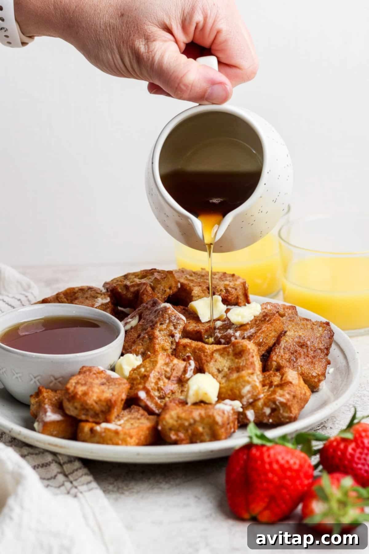 A hand pouring amber maple syrup over a plate of freshly made French toast cubes, glistening with butter and powdered sugar. A small bowl of syrup and fresh strawberries are nearby, with orange juice glasses in the background.