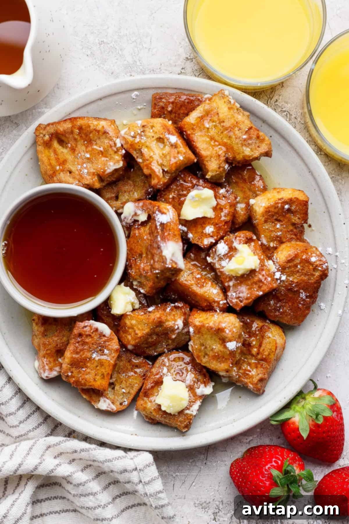 A vibrant plate of Air Fryer French toast bites, beautifully arranged with melting butter, a generous dusting of powdered sugar, and a side of maple syrup. Fresh strawberries and glasses of orange juice complete the inviting breakfast scene.