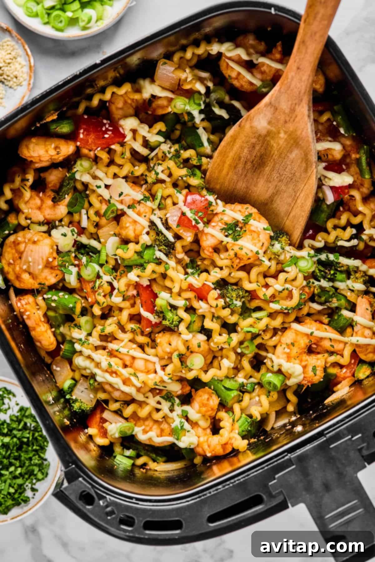 Crispy Air Fryer Shrimp & Veggies 2 A wooden spoon stirs a colorful mix of curly pasta, plump shrimp, crisp vegetables, and fresh herbs in an air fryer basket, with bowls of bright green onions and chopped herbs nearby.