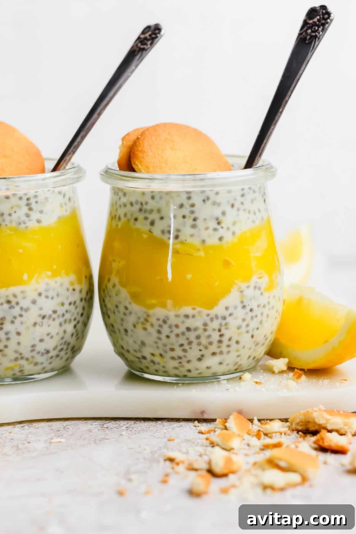 Two jars of chia pudding layered with lemon curd, each garnished with a cookie, placed on a marble surface with lemon wedges and cookie crumbs nearby.
