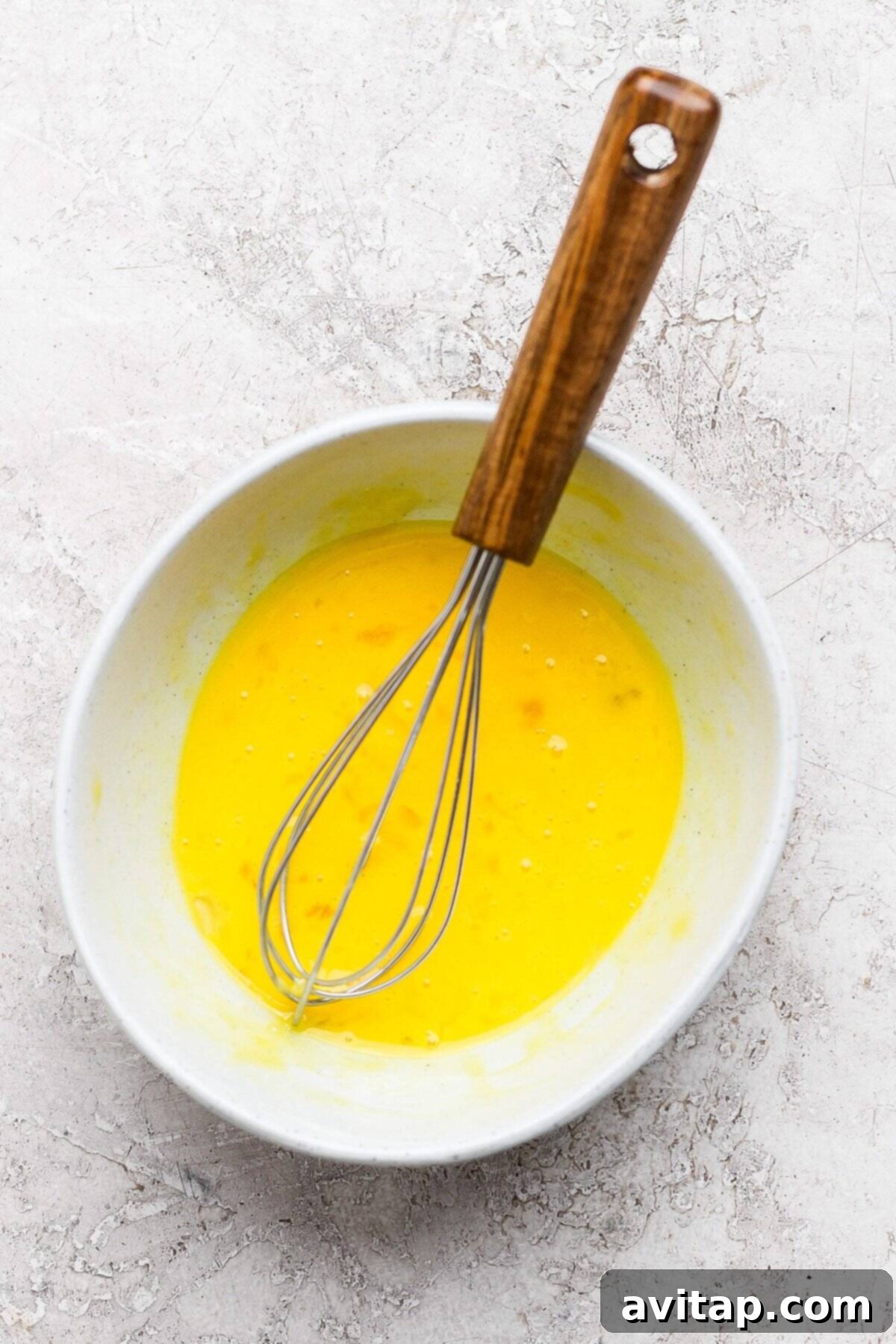 Whisk in a bowl of beaten eggs on a textured, light-colored surface.
