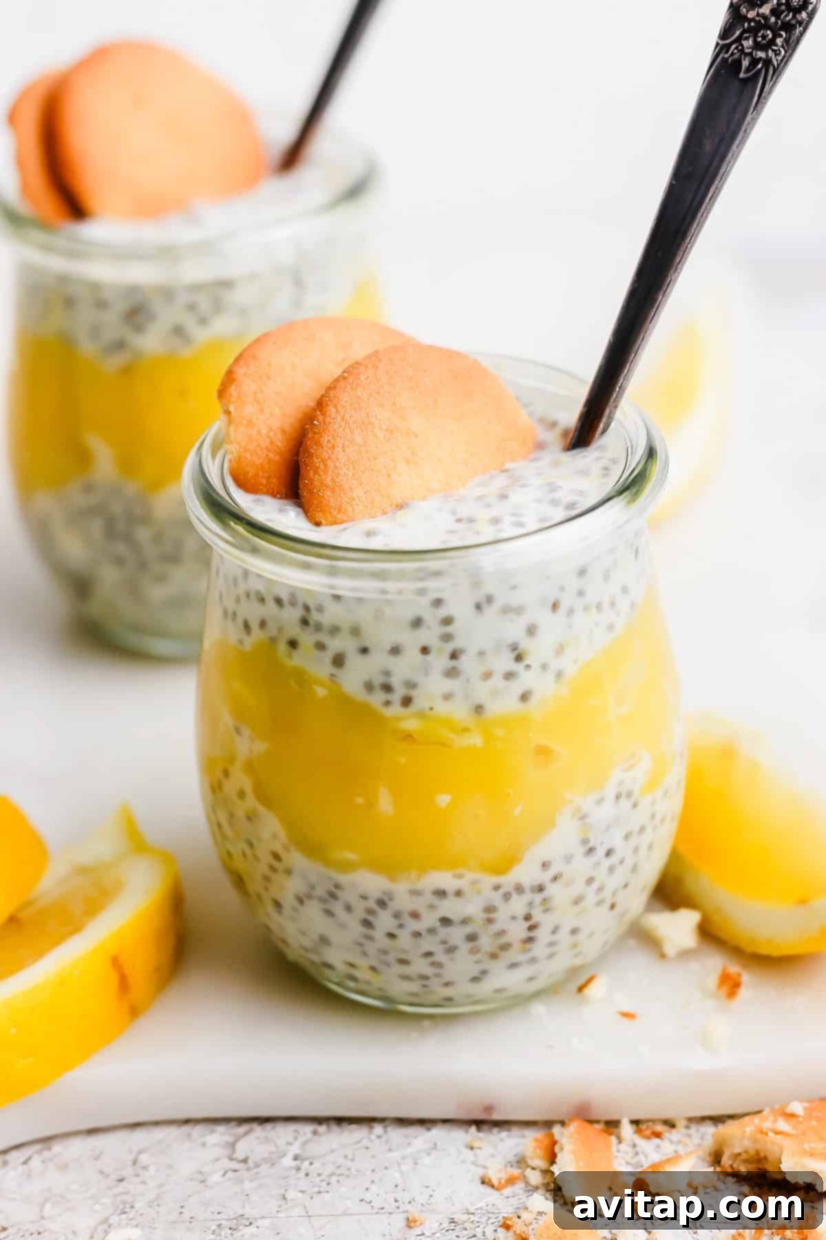 Protein Lemon Cream Dream Chia Pudding 2 Two clear glass jars filled with layers of creamy white chia pudding and vibrant yellow lemon curd, beautifully topped with two crisp Nilla Wafer cookies and a small spoon. The arrangement is artfully surrounded by fresh lemon slices and a scattering of cookie crumbs on a light, inviting surface.
