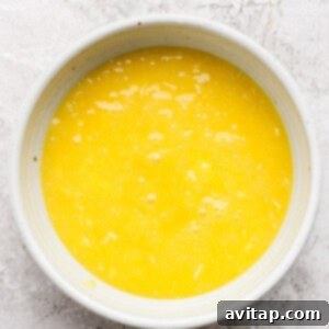 A white bowl filled with freshly cooked lemon curd, now covered with plastic wrap pressed directly onto its surface, cooling on a textured white surface.