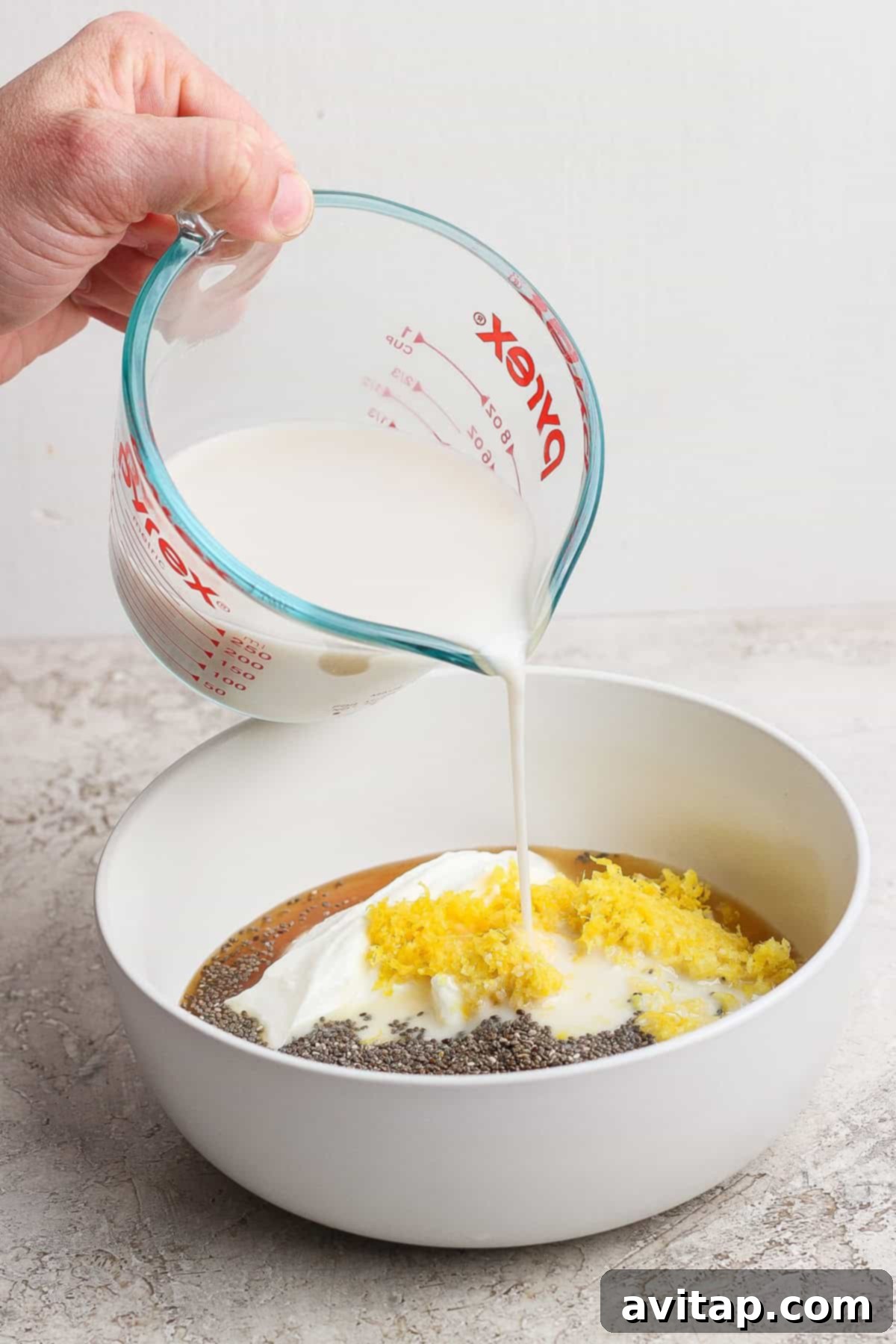 Protein Lemon Cream Dream Chia Pudding 3 A hand pouring unsweetened almond milk from a glass measuring cup into a large white mixing bowl. The bowl already contains blended 2% cottage cheese, tiny black chia seeds, bright yellow lemon zest, and a drizzle of honey, all ready to be thoroughly combined for the chia pudding base.