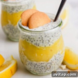 Close-up of a glass jar filled with luscious chia seed pudding and vibrant yellow lemon layers, beautifully topped with two crisp Nilla Wafer cookies and a small spoon. Fresh lemon slices are artfully arranged in the soft-focus background.