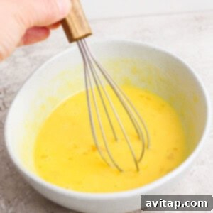 A hand expertly whisking yellow egg and egg yolk mixture in a gray bowl, preparing the base for the lemon curd.