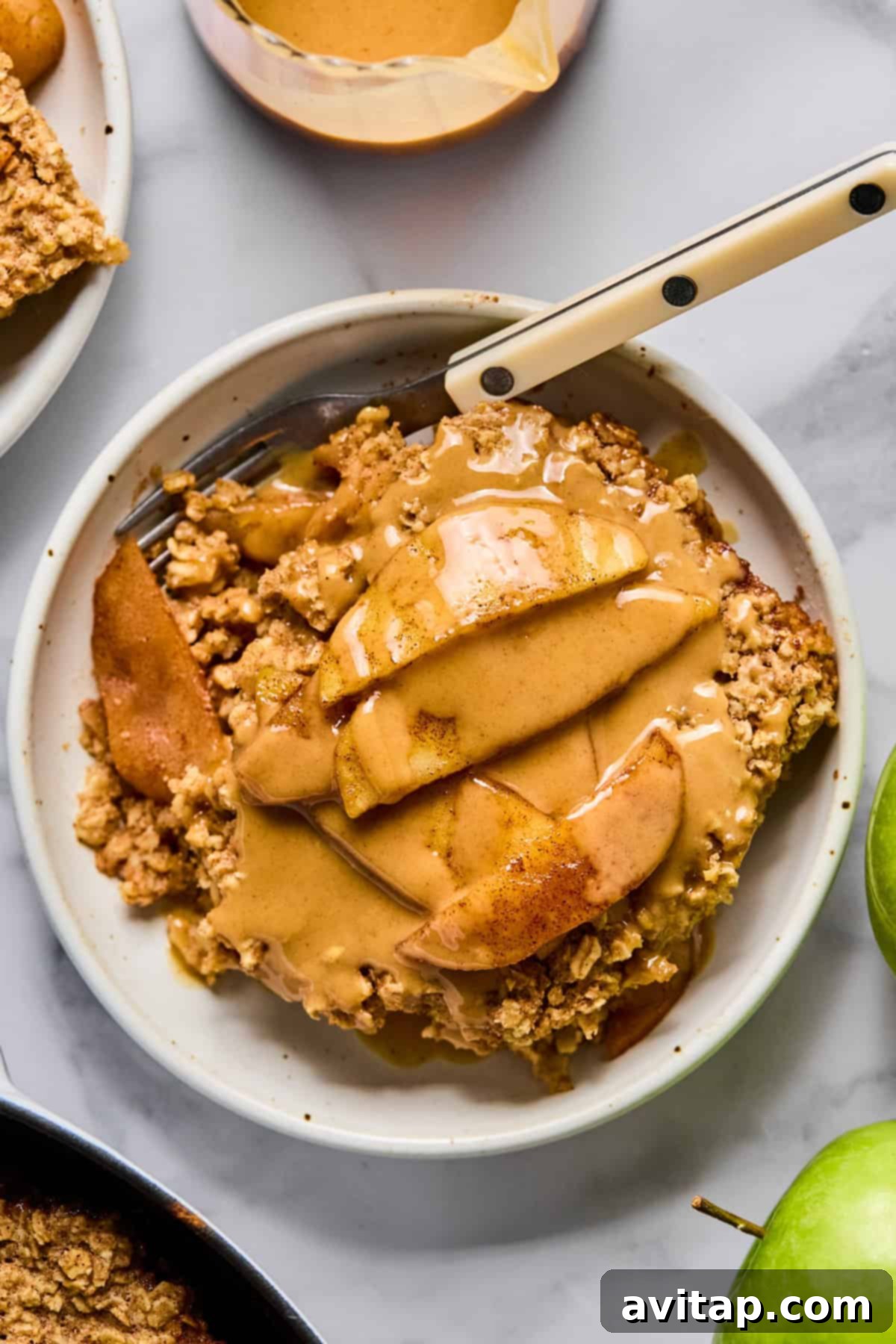 A slice of warm baked apple oatmeal, topped with tender sliced apples and a rich drizzle of peanut butter caramel sauce, elegantly presented on a white plate with a fork.