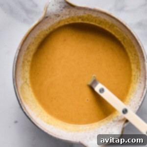 A ceramic bowl filled with a smooth, light brown liquid mixture of peanut butter caramel, with a white-handled spoon resting inside, placed on a elegant marble surface.