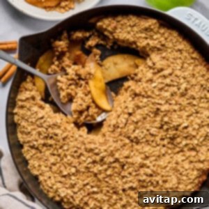 A cast iron skillet filled with baked apple crisp topped with oat crumble, with a serving scooped out onto a plate. Green apples and cinnamon sticks are nearby, ready for serving.