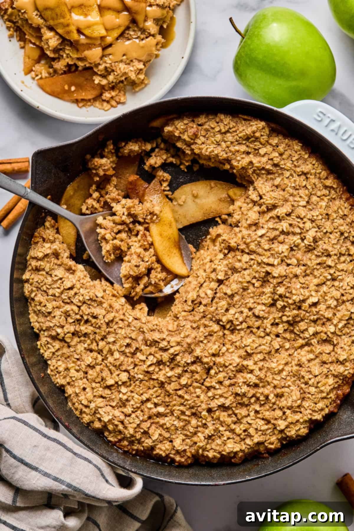 A warm cast iron skillet filled with freshly baked apple oatmeal, featuring a golden oat crumble topping. A portion has been scooped out onto a plate, with fresh green apples and cinnamon sticks artfully arranged nearby.