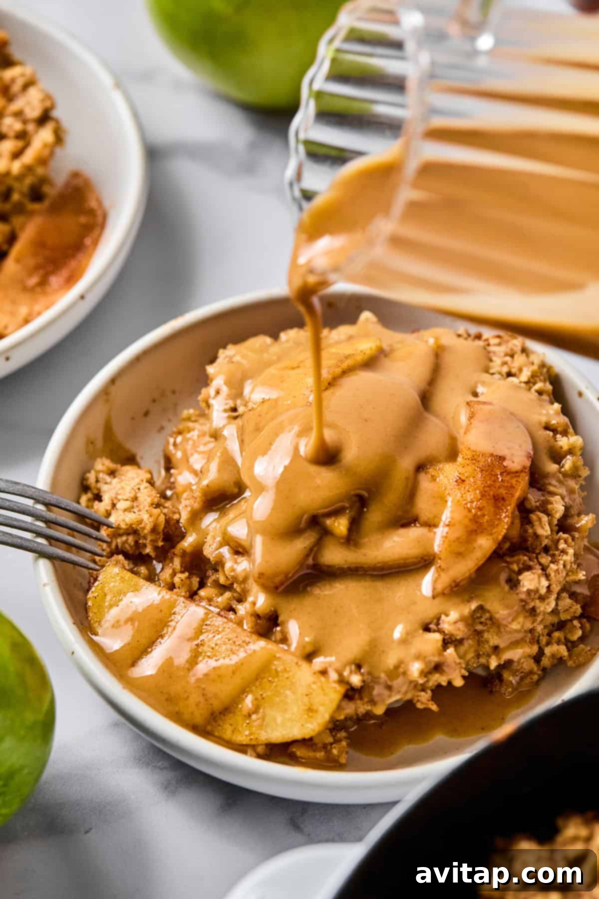 A beautiful bowl of baked apple oatmeal, generously drizzled with rich peanut butter caramel sauce from a glass pitcher, showcasing tender apple pieces and a fork resting gracefully on the side.