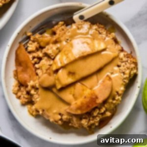 A bowl of apple crisp topped with sliced apples and a generous drizzle of peanut butter caramel, with a fork resting in the bowl, ready to be enjoyed.