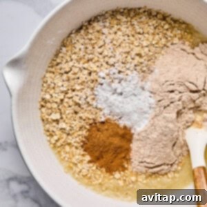 A mixing bowl containing oats, flour, baking powder, cinnamon, and a liquid ingredient with a spatula resting on the side, showcasing the oatmeal batter preparation.