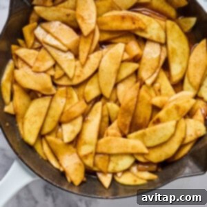 Sliced apples coated with cinnamon and sugar cooking in a cast iron skillet on a marble countertop, ready for the oat topping.