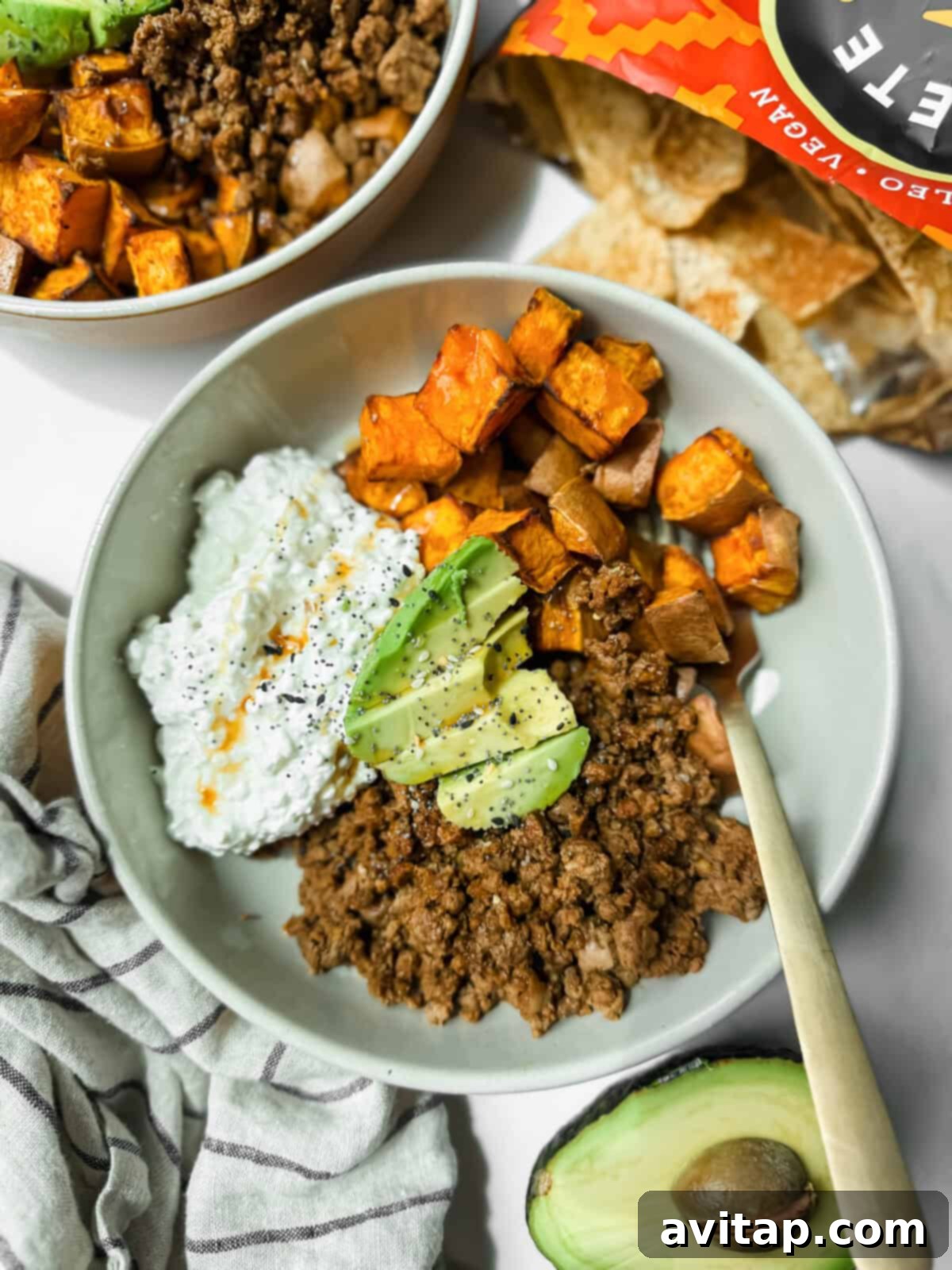 A vibrant bowl featuring seasoned ground beef, roasted sweet potato cubes, creamy cottage cheese, and perfectly sliced avocado. An open bag of tortilla chips and a halved avocado are visible nearby on a rustic wooden surface.