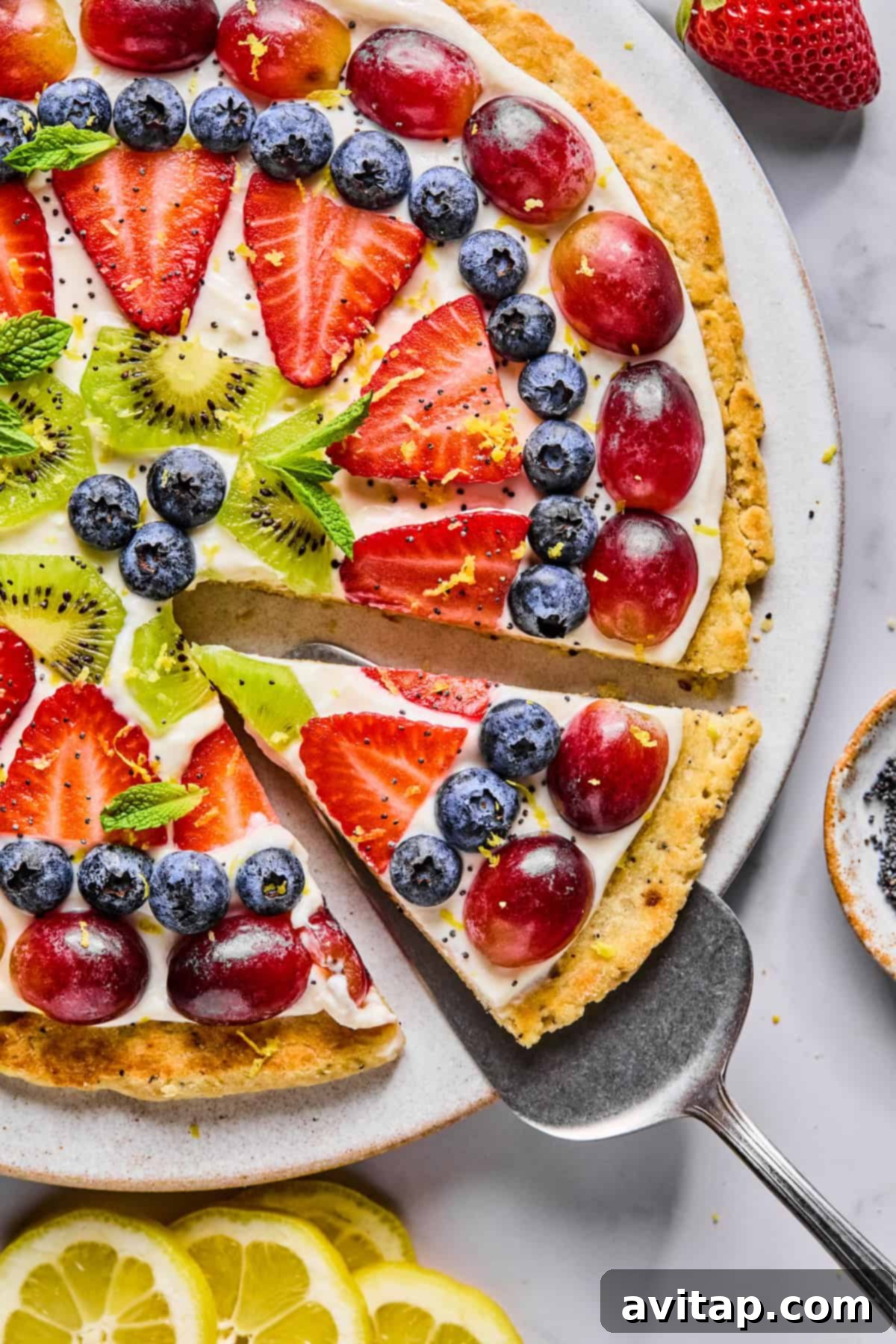 A fruit pizza topped with sliced strawberries, kiwi, blueberries, and red grapes on a creamy layer, with one slice being lifted from the whole.