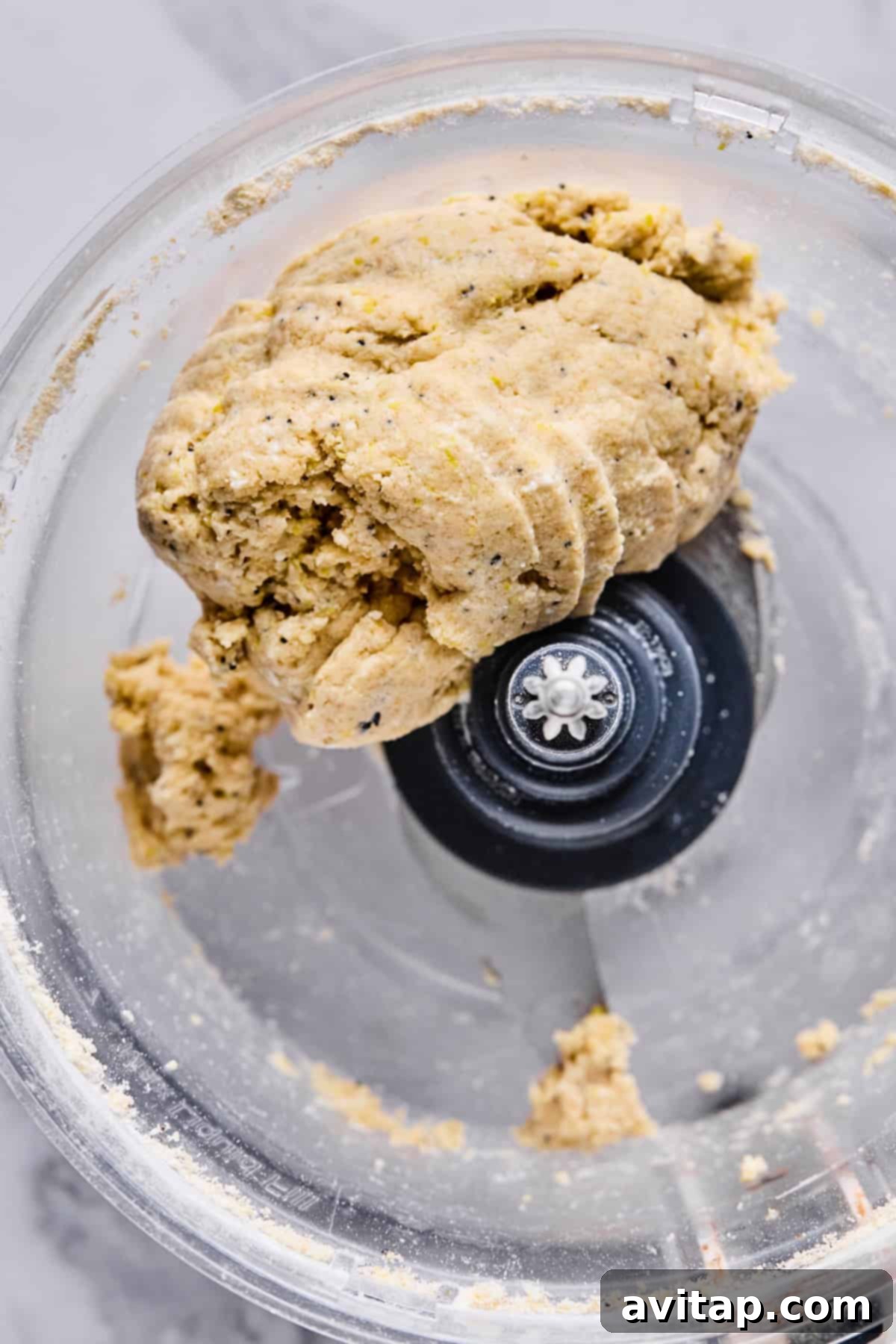 A close-up view of dough formed into a ball inside a food processor with a visible metal blade, indicating readiness for rolling.