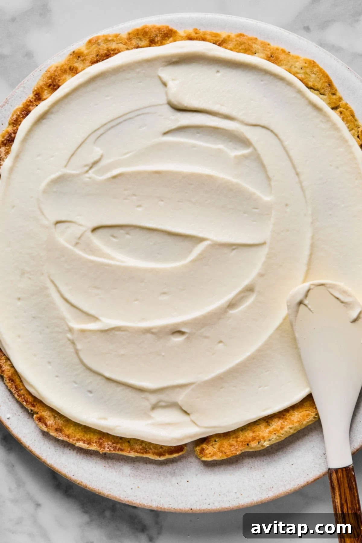 A partially frosted round cake on a plate, with a spatula spreading white frosting over the surface, showing the creamy texture of the Greek yogurt filling.