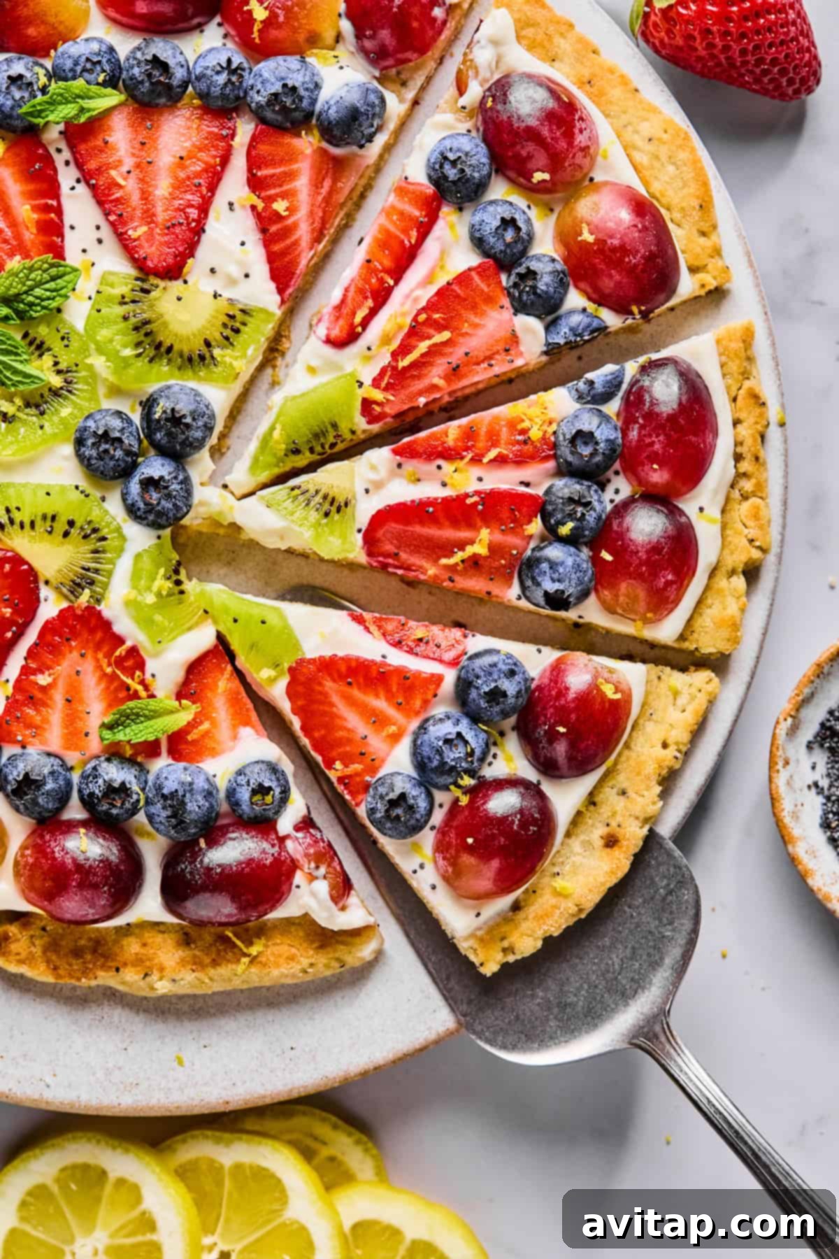 A fruit pizza topped with sliced strawberries, grapes, kiwi, and blueberries on a cream base, with a pie server lifting one slice from the plate, showcasing its vibrant appeal.