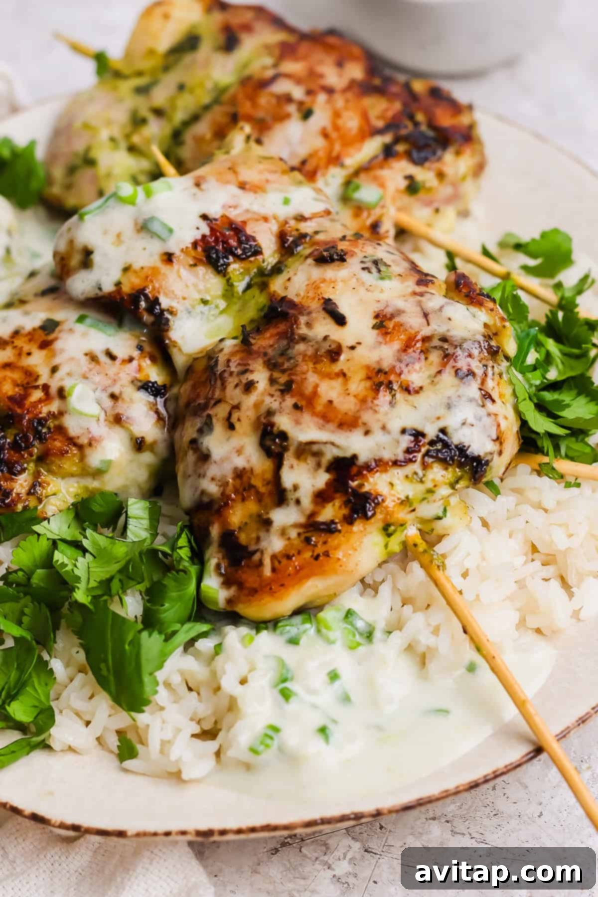 Three perfectly grilled chicken skewers, beautifully drizzled with creamy white coconut sauce, presented over a bed of fluffy white rice and garnished with fresh cilantro on a pristine white plate.