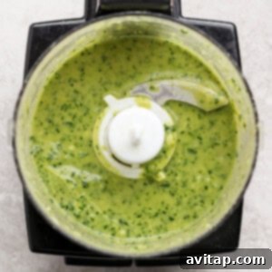 Overhead view of a food processor containing a smooth, bright green sauce or mixture with visible herbs on a light countertop.
