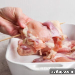 A hand holding a raw chicken piece on a wooden skewer over a white ceramic baking dish with more raw chicken pieces inside.