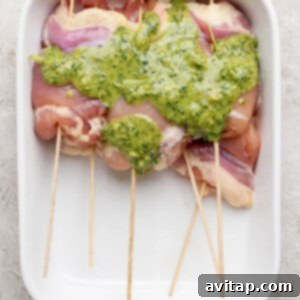 Raw chicken breasts skewered on wooden sticks in a white baking dish, topped with a green herb sauce, ready for marinating.