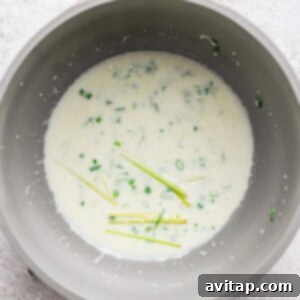 A saucepan containing a creamy white sauce with chopped green herbs and several stalks of lemongrass on a light textured surface, simmering gently.