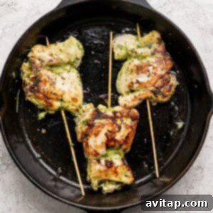 Three grilled, stuffed chicken thighs secured with wooden skewers are cooking in a black cast iron skillet on a light textured surface, sizzling.
