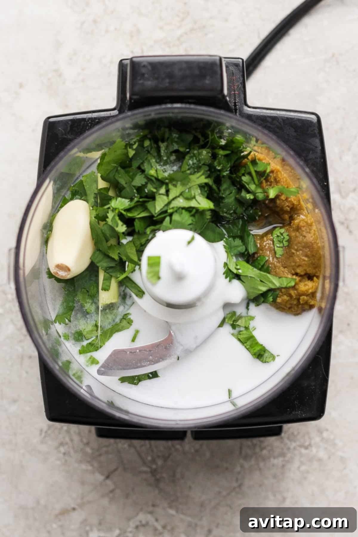 Overhead view of a food processor with coconut milk, fresh cilantro, garlic, ginger, and green curry paste, ready for blending into marinade.
