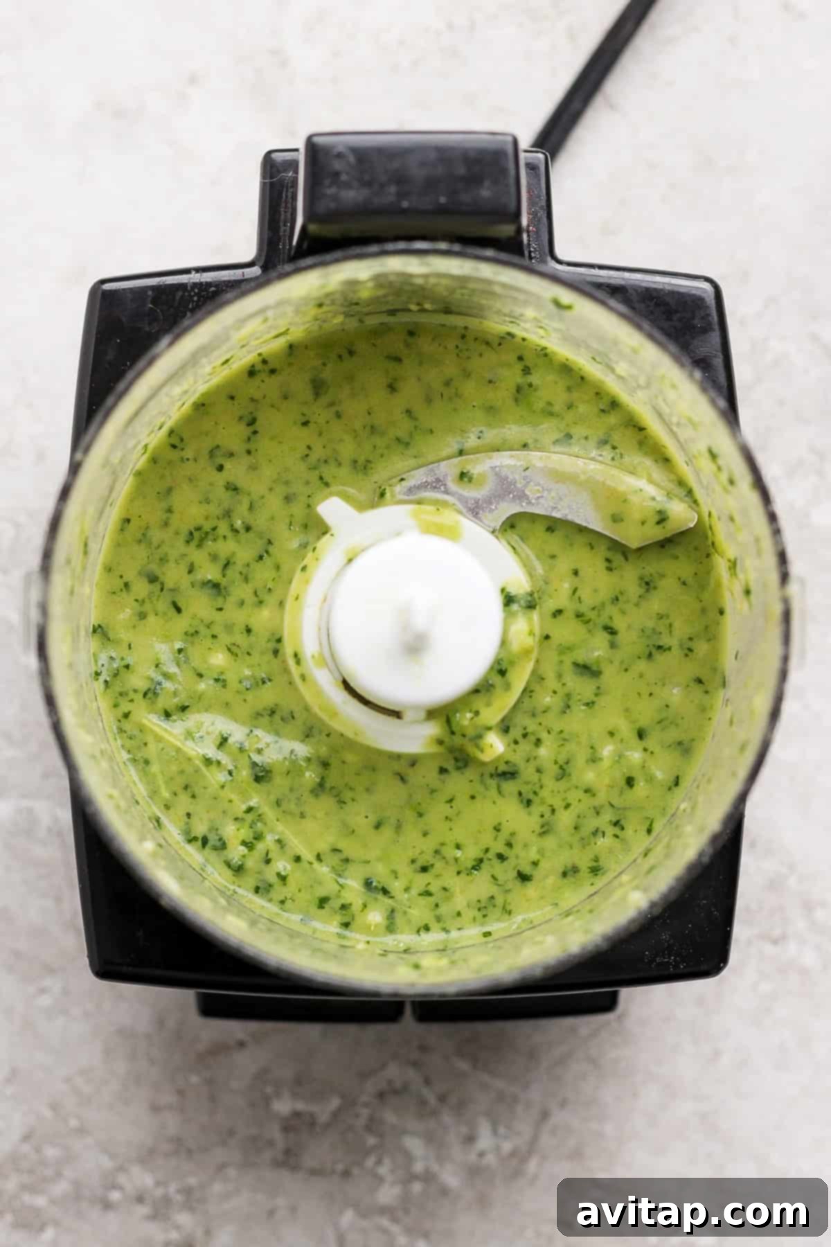 Overhead view of a food processor containing a smooth, bright green marinade mixture with visible herb flecks.