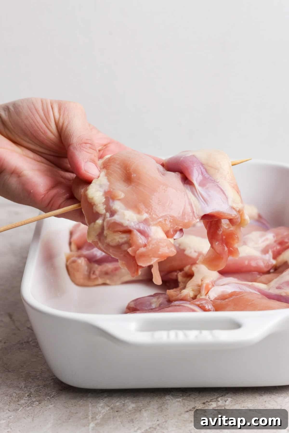A hand expertly threading a raw chicken piece onto a wooden skewer over a baking dish filled with more raw chicken.
