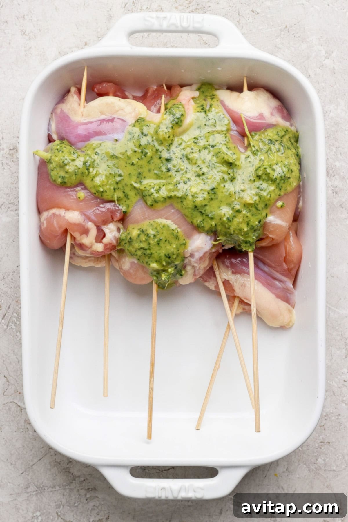 Raw chicken breasts skewered on wooden sticks, arranged in a white baking dish and coated in a lush green herb sauce, ready for marination.
